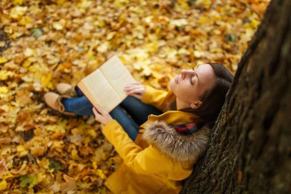 Piękny szczęśliwy uśmiechający się Brunet Kobieta w żółtym fartuchu i dżinsy, siedząc pod drzewem klonowym czerwony książkę w Upadek miasta park na ciepły dzień. Autumn golden odchodzi. Koncepcja czytania. Widok z góry. — Zdjęcie stockowe