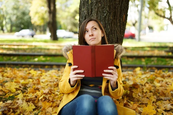 Piękny szczęśliwy uśmiechający się Brunet Kobieta w żółtym fartuchu i dżinsy, siedząc pod drzewem klonowym czerwony książkę w Upadek miasta park na ciepły dzień. Autumn golden odchodzi. Koncepcja czytania — Zdjęcie stockowe