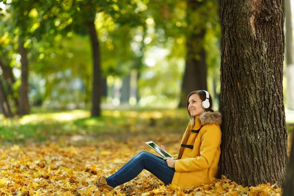 黄色のコートとジーンズ座って、そして暖かい日に彼女の手でタブレットと秋都市公園におけるヘッドフォンとツリーの下で音楽を聴いて笑顔茶色の髪の女性。黄金の秋の紅葉. — ストック写真