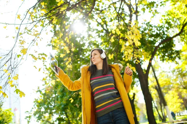 A beautiful happy cheerful brown-haired woman in a yellow coat and striped longsleeve rejoices with a tablet in her hands and white headphones in fall city park on a warm day. Autumn golden leaves.