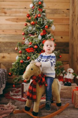 Mutlu sevimli küçük oğlu üzerinde sallanan at bir oyuncak ile Noel ağacı, giyinmiş kazak ve kot evde dekore edilmiş yeni yıl odasında. Çocuk iyi bir ruh hali. Yaşam tarzı, Aile ve tatil 2018 kavramı