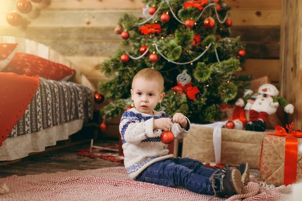 Happy cute little baby boy dressed in sweater and jeans decorating Christmas tree with toys in wooden room at home. Child with good mood. New Year. Lifestyle, family and holiday 2018 concept — Stock Photo, Image