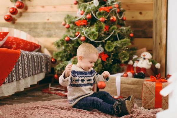 Feliz lindo niño vestido con suéter y jeans decorando el árbol de Navidad con juguetes en la habitación de madera en casa. Niño con buen humor. Año Nuevo. Estilo de vida, familia y vacaciones 2018 concepto — Foto de Stock