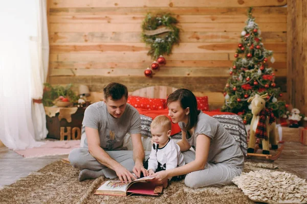 Μικρά χαρούμενα ευτυχείς γονείς με χαριτωμένο μικρό παιδί γιος στο πάτωμα ανάγνωση παραμυθιού στο βιβλίο κοντά στο χριστουγεννιάτικο δέντρο διακόσμηση νέο έτος ξύλινο δωμάτιο στο σπίτι. Τρόπο ζωής οικογένεια και διακοπές έννοια 2018 — Φωτογραφία Αρχείου