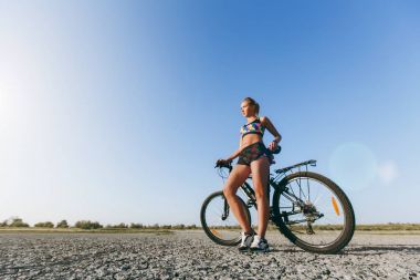 Güçlü bir sarışın kadın çok renkli elbiseli yakınındaki bir çöl bölgesinde bir bisiklet standları ve güneşe benziyor. Fitness kavramı. Mavi gökyüzü arka plan
