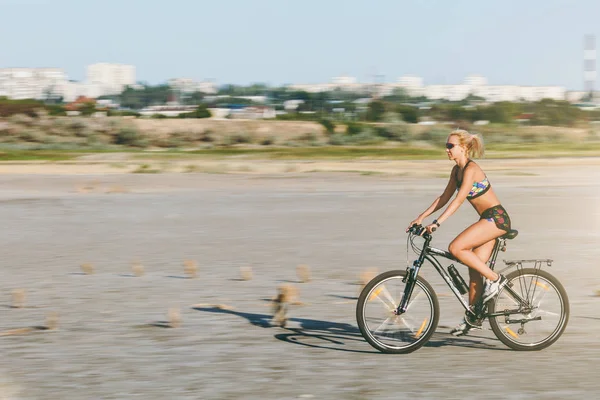 Uma mulher loira esportiva em um terno colorido anda de bicicleta em velocidade rápida em uma área deserta em um dia ensolarado de verão. Conceito de aptidão. Plano de fundo — Fotografia de Stock