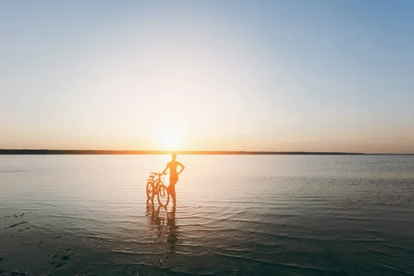 Una donna bionda forte in un abito colorato si trova vicino alla bicicletta in acqua al tramonto in una calda giornata estiva. Concetto fitness. Sfondo cielo — Foto Stock