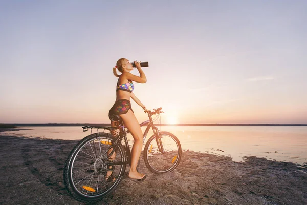 A strong blonde woman in a colorful suit sits on the bicycle, drinks water from a black bottle in a desert area near the water. Fitness concept. Blue sky background — Stock Photo, Image