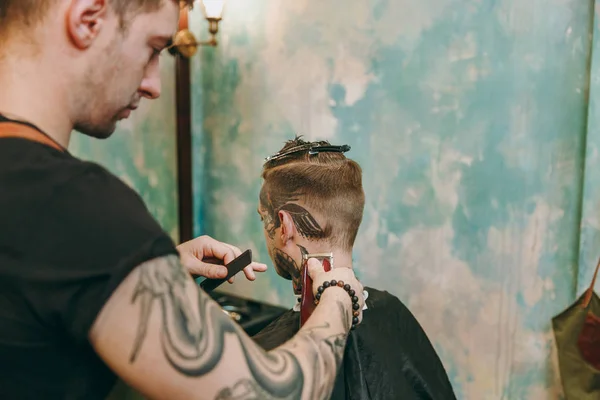 Tiro de perto de um homem a cortar o cabelo na barbearia. Cabeleireiro masculino em tatuagens servindo cliente . — Fotografia de Stock