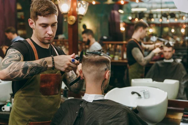 A man getting trendy haircut at barber shop. Male hairstylist in tattoos serving client. — Stock Photo, Image