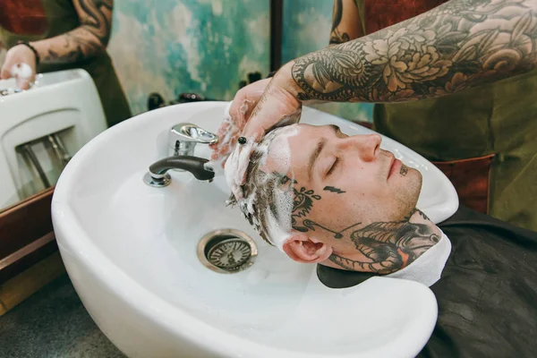 Крупный план человека, стригущегося в парикмахерской. Мужчина-парикмахер в татуировках обслуживающий клиента, моющая голова — стоковое фото