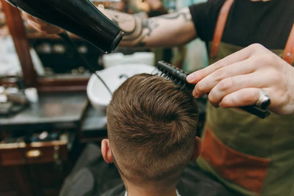 Крупним планом чоловік стає модною стрижкою в перукарні. Чоловічий перукар в татуюваннях, що обслуговують клієнта, сушка волосся з фен — стокове фото