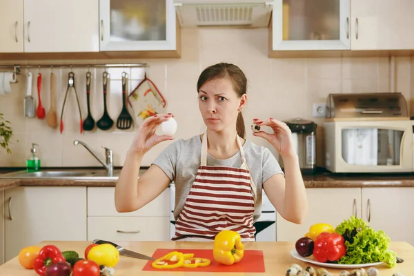 Eine junge attraktive, nachdenkliche Frau in Schürze wählt in der Küche zwischen Hühner- und Wachteleiern. Diätkonzept. Gesunder Lebensstil. Kochen zu Hause. Essen zubereiten. — Stockfoto