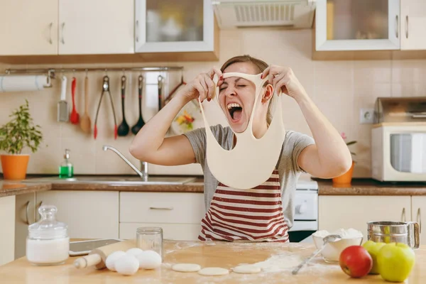 Uma jovem mulher alegre e sorridente engraçada coloca em uma massa com buracos no rosto e se diverte na cozinha. A cozinhar para casa. Preparar alimentos . — Fotografia de Stock