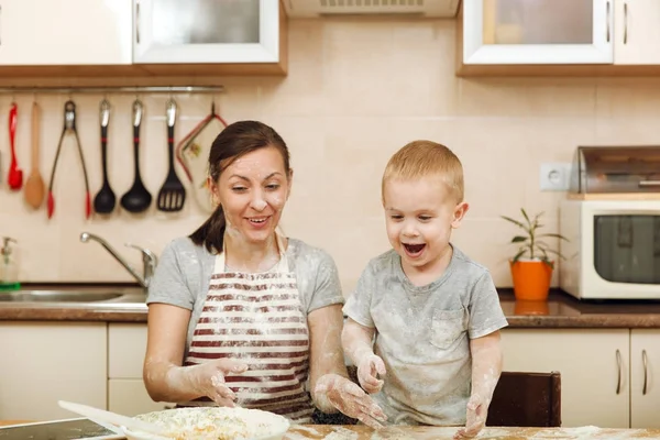 Little kid boy helps mother to cook Christmas ginger biscuit in light kitchen with tablet on the table. Happy family mom 30-35 years and child 2-3 have fun and throw flour home. Relationship concept — Stock Photo, Image