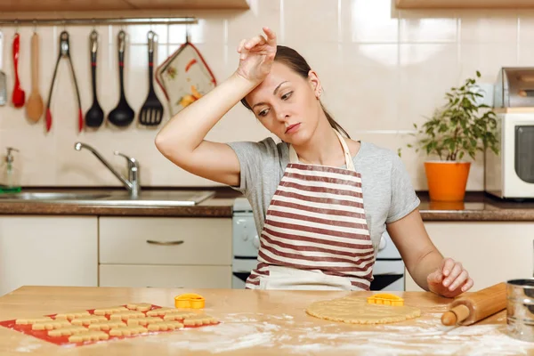 Een jonge mooie moe vrouw 30-35 jaar in schort zittend aan een tafel met deeg en deegroller en gaan te bereiden een kerst gember taarten in de lichte keuken. Home koken. Eten bereiden. — Stockfoto