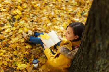 Güzel bir mutlu kahverengi saçlı kadın sarı ceket ve kot fincan çay veya kahve ve kırmızı kitap sonbahar şehir parkı ile akçaağaç ağacın altında oturuyor. Altın yaprak. Okuma kavramı. Üstten Görünüm.