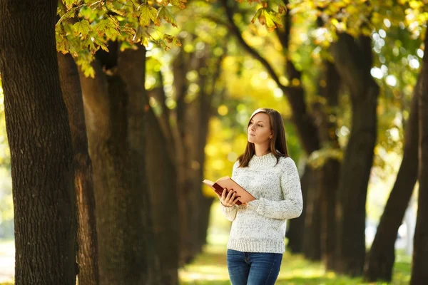 A beautiful happy smiling brown-haired woman in white sweater standing with a red book in fall city park on a warm day. Autumn golden leaves. Reading concept.