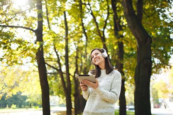 A beautiful happy cheerful brown-haired woman in white sweater with a tablet listening music in the white headphones in fall park on a warm day. Autumn in the city.