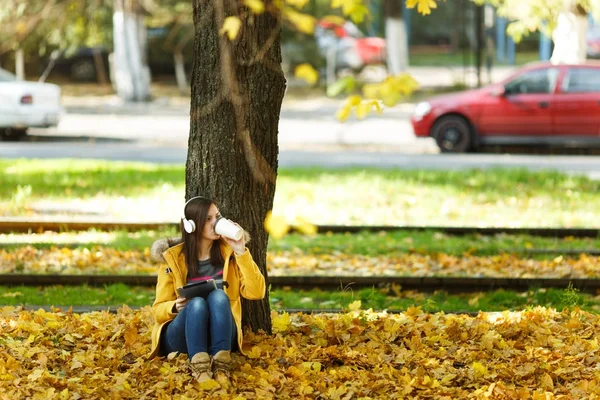 Uma mulher de casaco amarelo e jeans sentada com uma xícara de café ou chá e ouvindo música debaixo de uma árvore com um tablet nas mãos e fones de ouvido no parque da cidade de outono em um dia quente. Outono folhas douradas . — Fotografia de Stock