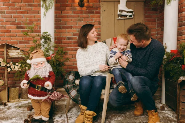 Νέοι ευτυχείς γονείς με ένα χαριτωμένο μικρό παιδί αγόρι ντυμένος με πουλόβερ που κάθεται στην καρέκλα με Santa στο διακοσμημένο δωμάτιο στο σπίτι με το νέο έτος. Χριστούγεννα καλή διάθεση. Τρόπου ζωής, οικογένεια και διακοπές έννοια 2018 — Φωτογραφία Αρχείου