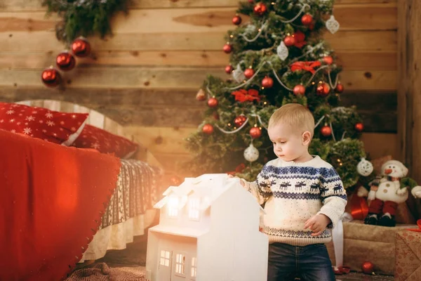 Feliz niño lindo vestido con suéter sentado en el árbol de Navidad con una casa blanca de juguete mágico en la habitación de madera en casa. Buen humor para niños. Año Nuevo. Estilo de vida, familia y vacaciones 2018 concepto — Foto de Stock