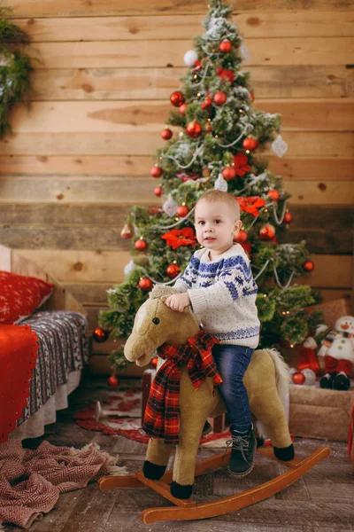 Ευτυχισμένος χαριτωμένο μικρό μωρό αγόρι κουνιστό άλογο στο χριστουγεννιάτικο δέντρο με ένα παιχνίδι, ντυμένος με πουλόβερ και τζιν στο διακοσμημένο δωμάτιο στο σπίτι με το νέο έτος. Παιδί καλή διάθεση. Τρόπου ζωής, οικογένεια και διακοπές έννοια 2018 — Φωτογραφία Αρχείου
