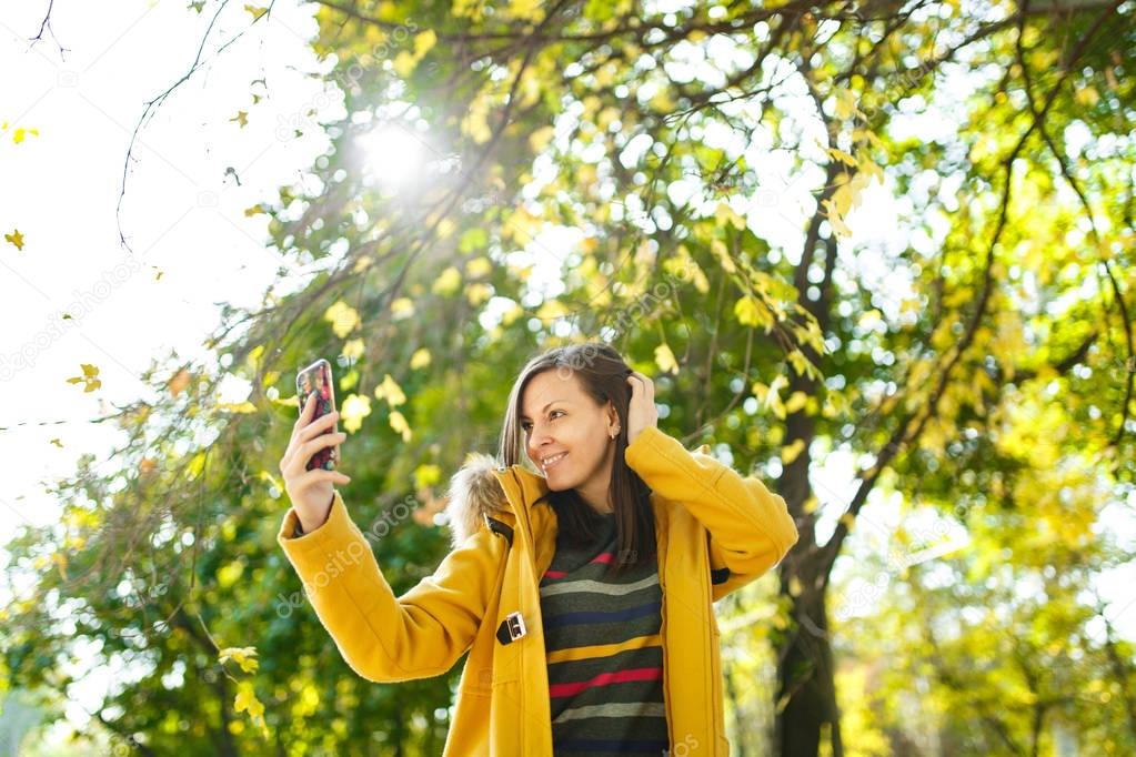 A beautiful happy cheerful brown-haired woman in a yellow coat and striped longsleeve doing selfie on mobile phone in fall city park on a warm day. Autumn golden leaves.