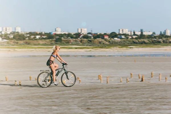 Uma mulher loira esportiva em um terno colorido anda de bicicleta em uma área deserta em um dia ensolarado de verão. Conceito de aptidão. Céu azul fundo — Fotografia de Stock