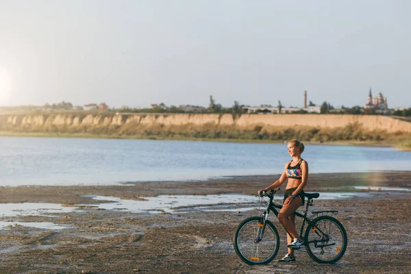 A strong blonde woman in a colorful suit sits on a bicycle in a desert area near the water and looks at the sun. Fitness concept. Blue sky background — Stock Photo, Image