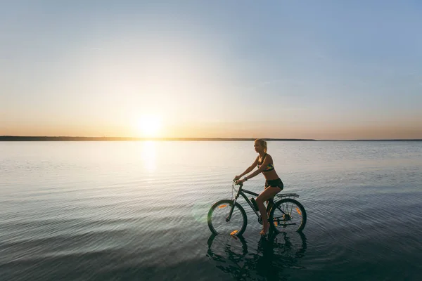 Eine kräftige blonde Frau im bunten Anzug sitzt bei Sonnenuntergang an einem warmen Sommertag auf dem Fahrrad im Wasser. Fitness-Konzept. Himmel Hintergrund — Stockfoto