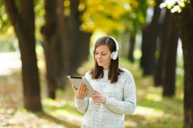 Güzel bir mutlu neşeli kahverengi saçlı Bayan Beyaz kazaklı bir tablet sonbahar Park'ta sıcak bir gün beyaz kulaklık müzik dinleme. Şehirde sonbahar.