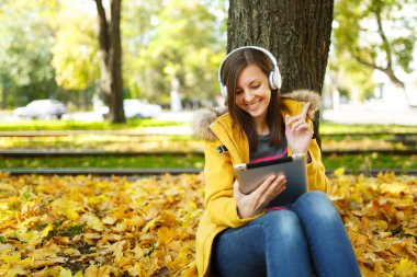 Kahverengi saçlı gülümseyen kadın sarı ceket ve kot oturan ve sıcak bir günde bir tablet onun elinde ve sonbahar Şehir Parkı kulaklık ile bir ağacın altında müzik dinlemek. Altın yaprak.