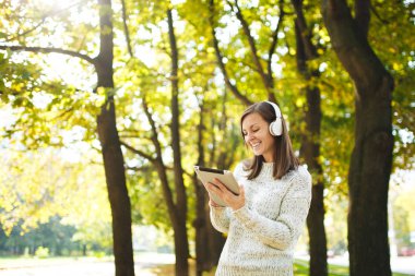 Güzel bir mutlu neşeli kahverengi saçlı Bayan Beyaz kazaklı bir tablet sonbahar Park'ta sıcak bir gün beyaz kulaklık müzik dinleme. Şehirde sonbahar.