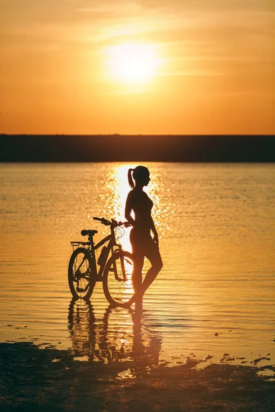 Silhouette of a sporty girl in a suit standing near a bicycle in the water at sunset on a warm summer day. Fitness concept. Sky background — Stock Photo, Image