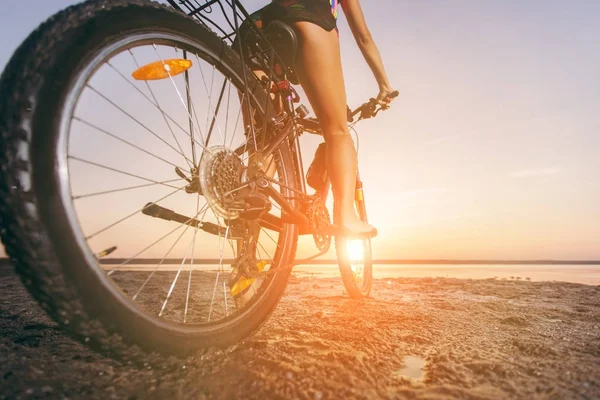 Kobieta w garnitur wielobarwny siedzi na rowerze w okolicy pustyni w pobliżu wody. Koncepcja fitness. Widok widok z tyłu i z dołu. Szczelnie-do góry — Zdjęcie stockowe