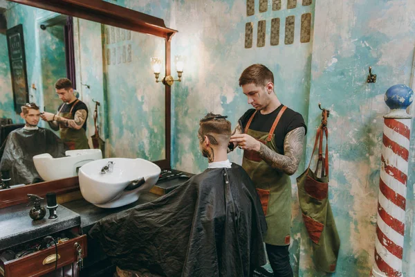 A man getting trendy haircut at barber shop. Male hairstylist in tattoos serving client. — Stock Photo, Image