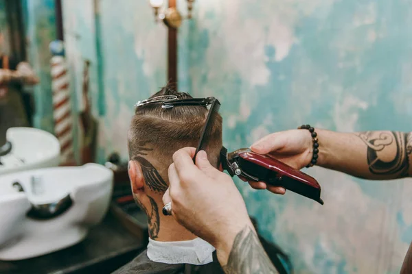 Tiro de perto de um homem a cortar o cabelo na barbearia. Cabeleireiro masculino em tatuagens servindo cliente . — Fotografia de Stock