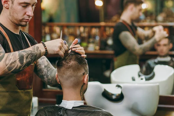 Close up shot of man getting trendy haircut at barber shop. Male hairstylist in tattoos serving client. — Stock Photo, Image