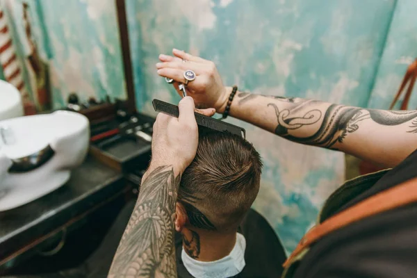 Tiro de perto de um homem a cortar o cabelo na barbearia. Cabeleireiro masculino em tatuagens servindo cliente . — Fotografia de Stock