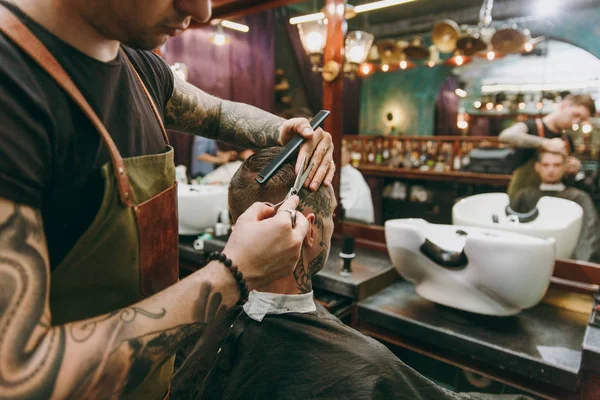 Close up shot of man getting trendy haircut at barber shop. Male hairstylist in tattoos serving client. — Stock Photo, Image