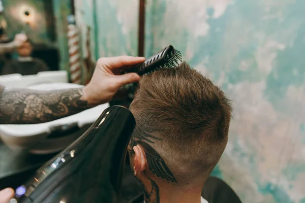 Close up shot of man getting trendy haircut at barber shop. Male hairstylist in tattoos serving client, drying hair with a hairdryer — Stock Photo, Image