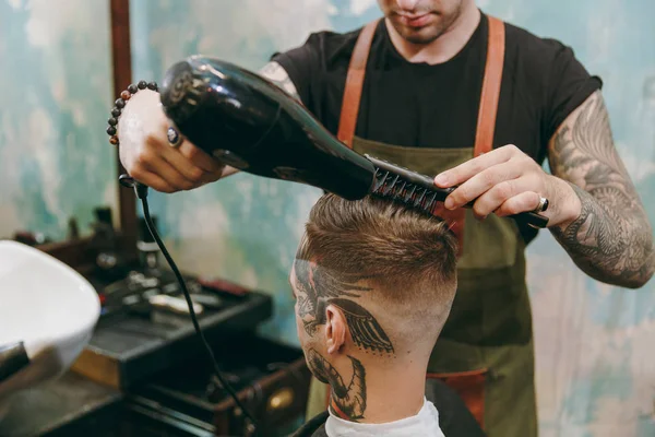 Close up shot of man getting trendy haircut at barber shop. Male hairstylist in tattoos serving client, drying hair with a hairdryer — Stock Photo, Image