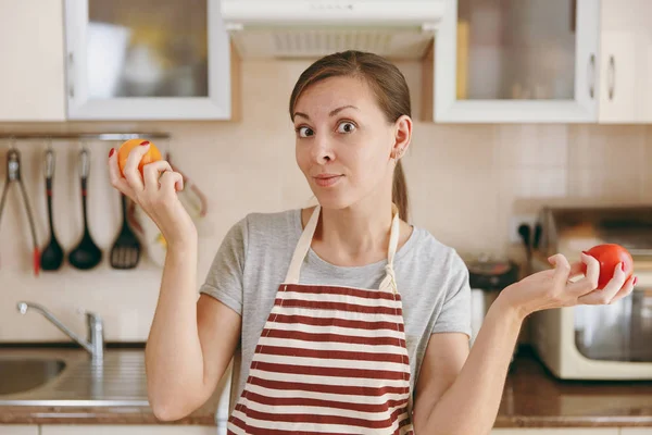 Eine junge attraktive, nachdenkliche Frau in Schürze entscheidet sich für eine rote oder gelbe Tomate in der Küche. Diätkonzept. Gesunder Lebensstil. Kochen zu Hause. Essen zubereiten. — Stockfoto