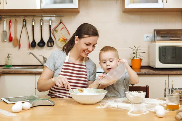 Little kid boy helps mother to cook Christmas ginger biscuit in light kitchen with tablet on the table. Happy family mom 30-35 years and child 2-3 in weekend morning at home. Relationship concept