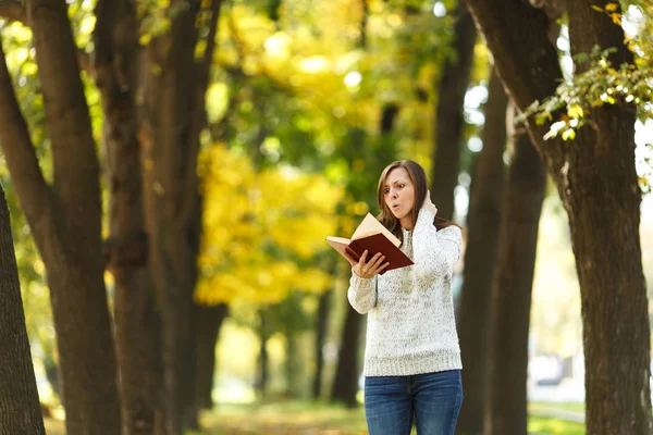 A beautiful happy smiling brown-haired woman in white sweater standing with a red book in fall city park on a warm day. Autumn golden leaves. Reading concept.