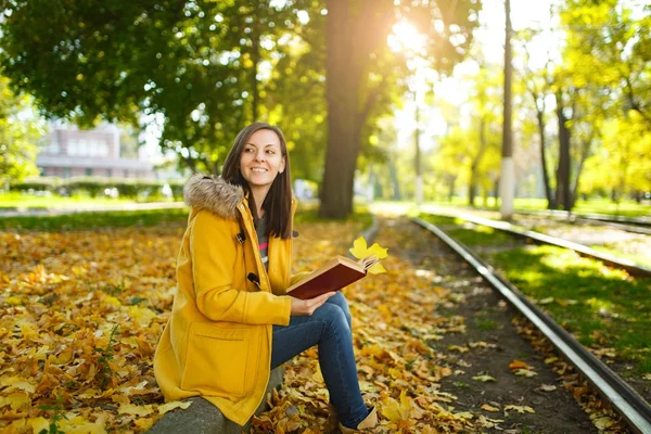 Piękne kobiety Brunet szczęśliwy w żółtym fartuchu i dżinsy samodzielnie znajduje się w parku w pobliżu torów tramwajowych i czyta książkę w ciepły dzień jesieni. Żółty jesienny liść. — Zdjęcie stockowe