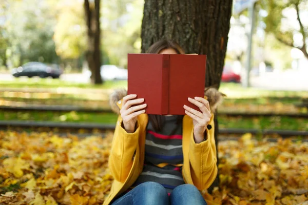 Piękny szczęśliwy uśmiechający się Brunet Kobieta w żółtym fartuchu i dżinsy, siedząc pod drzewem klonowym i chowając się za czerwone książki w Upadek miasta park na ciepły dzień. Autumn golden odchodzi. Koncepcja czytania — Zdjęcie stockowe