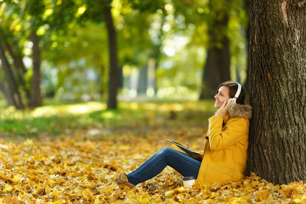 A woman in yellow coat and jeans sitting with cup of coffee or tea and listening to music under a tree with a tablet in her hands and headphones in fall city park on a warm day. Autumn golden leaves.
