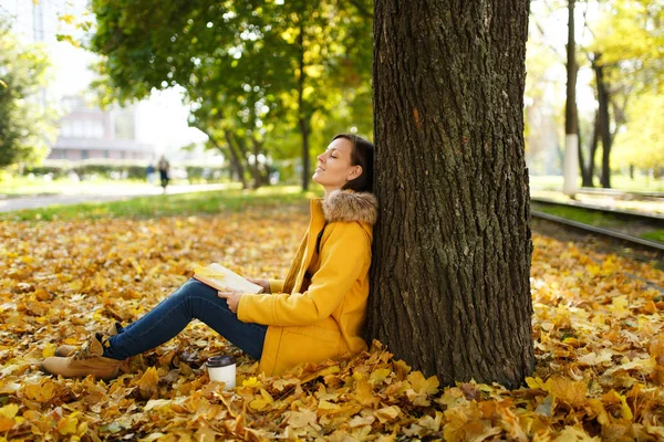 Piękna kobieta szczęśliwa Brunet w żółtym fartuchu i dżinsy, siedząc pod drzewem klonowym filiżankę herbaty lub kawy i czerwone książki w Upadek miasta park. Autumn golden odchodzi. Koncepcja czytania. — Zdjęcie stockowe